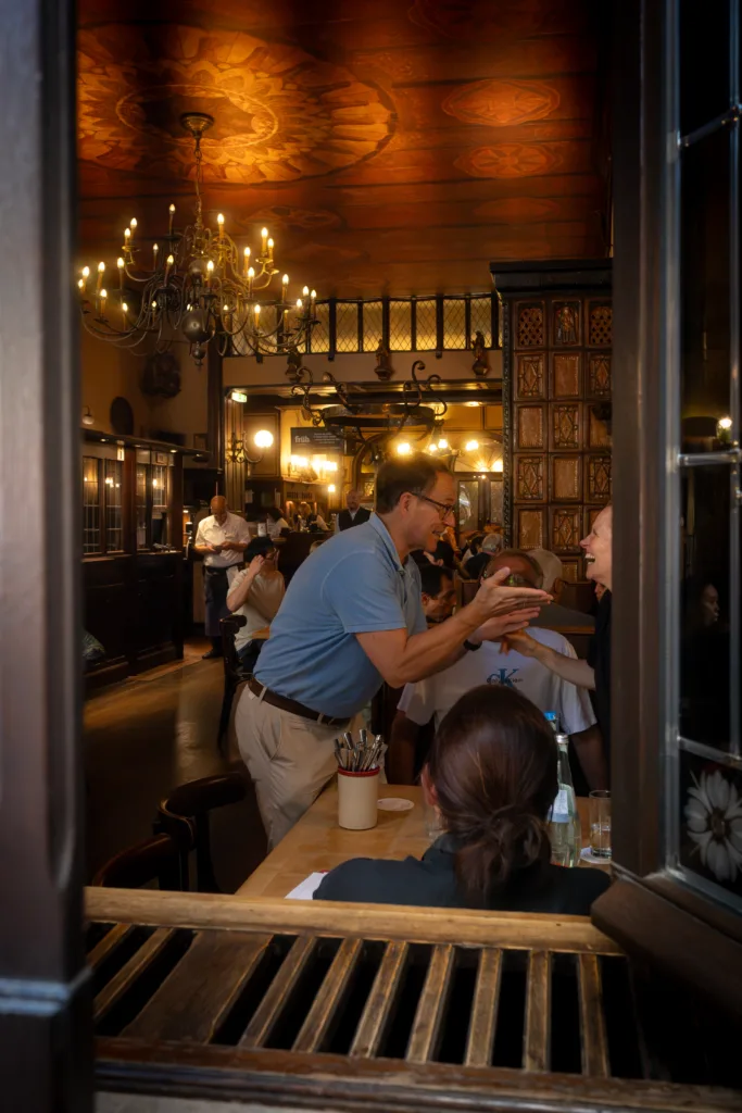 Two people greeting each other, laughing, in a Cologne brewery pub, chandelier and painted wooden ceiling behind