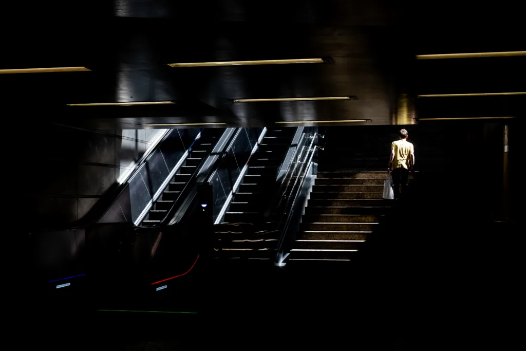 Person in a yellow shirt climbing the stairs in a dark underground station, escalators and strip lights alongside
