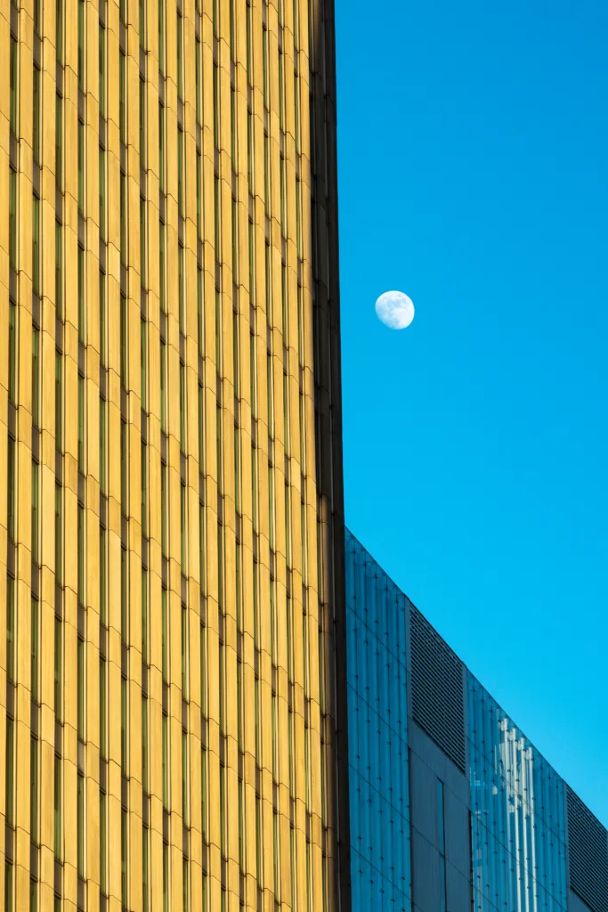 The moon in a deep blue daytime sky, in the gap between a golden ribbed facade and a dark blue-grey one