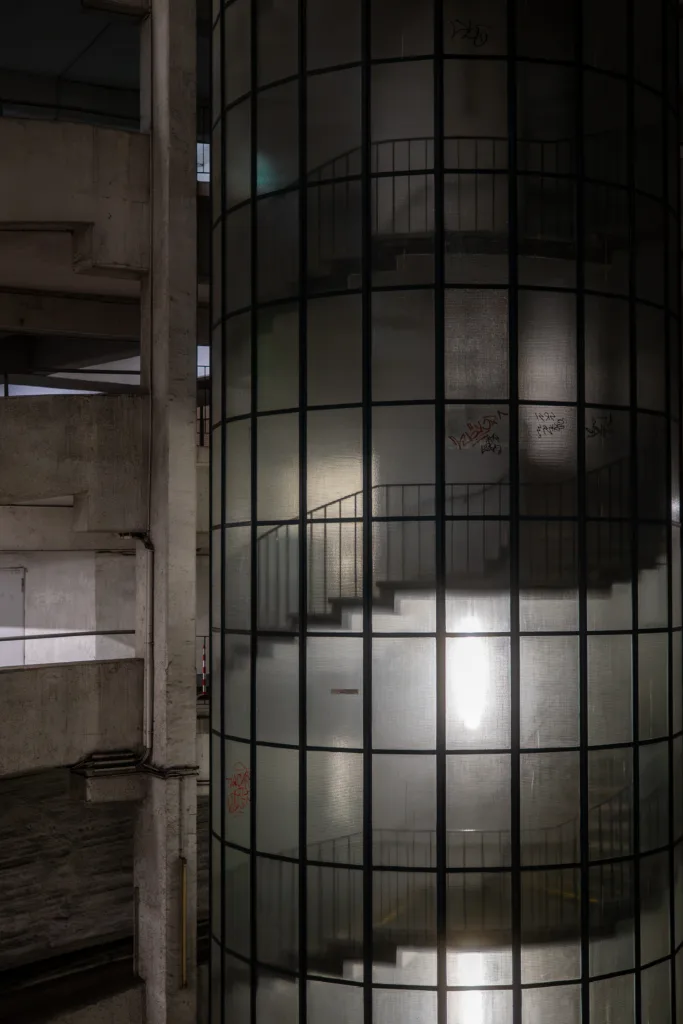 Cylindrical glass stairwell tower in a concrete car park at night, the staircase showing as a silhouette through frosted glass