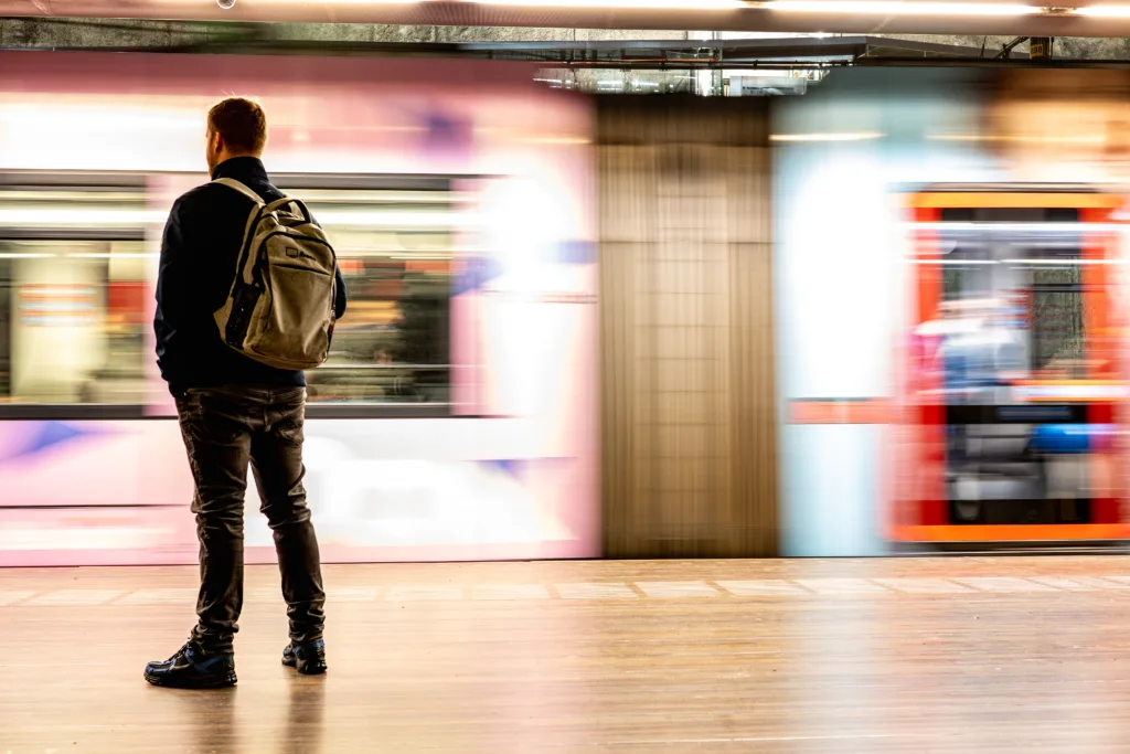 Person with a backpack standing still on an underground platform while two trains pass in blurred motion