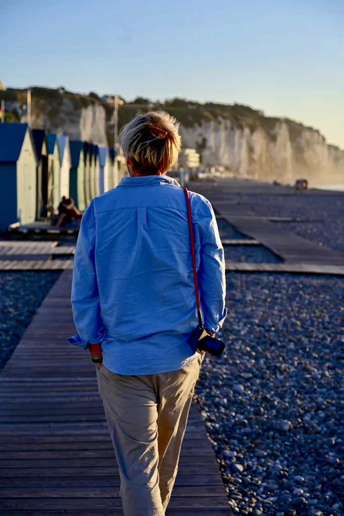 Photographer walking away along a boardwalk past beach huts, chalk cliffs and a pebble beach in the evening light